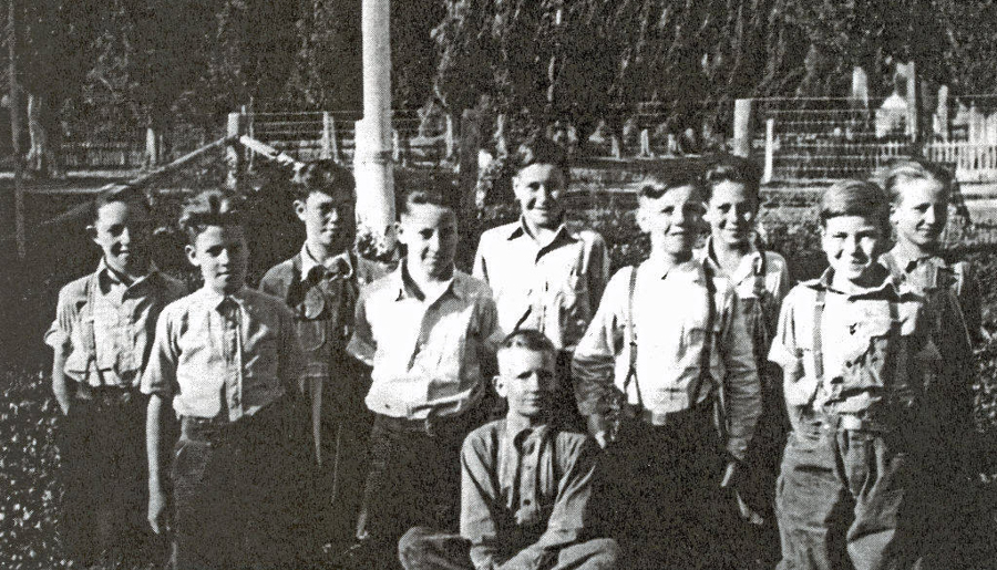 A group of ten boys, with eight standing and two sitting in front, are posed outdoors. They are dressed in shirts and trousers with suspenders visible on some. Trees and fencing are in the background.