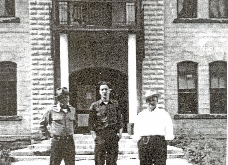 Three men standing in front of a building with a stone facade and columns. The man on the left wears a shirt and hat, the man in the middle is in dark clothing, and the man on the right wears a light shirt with a hat. There are steps leading up to the entrance, and windows are visible on the building.