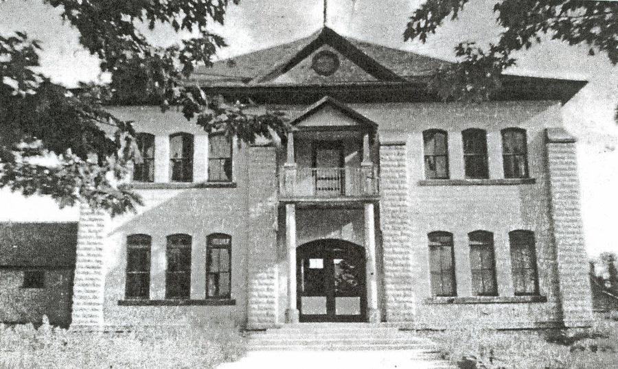 A two-story, symmetrical brick building is shown with a central entrance featuring a small porch and steps leading up to double doors. The building has multiple tall, narrow windows on both floors. Trees partially frame the view of the building.