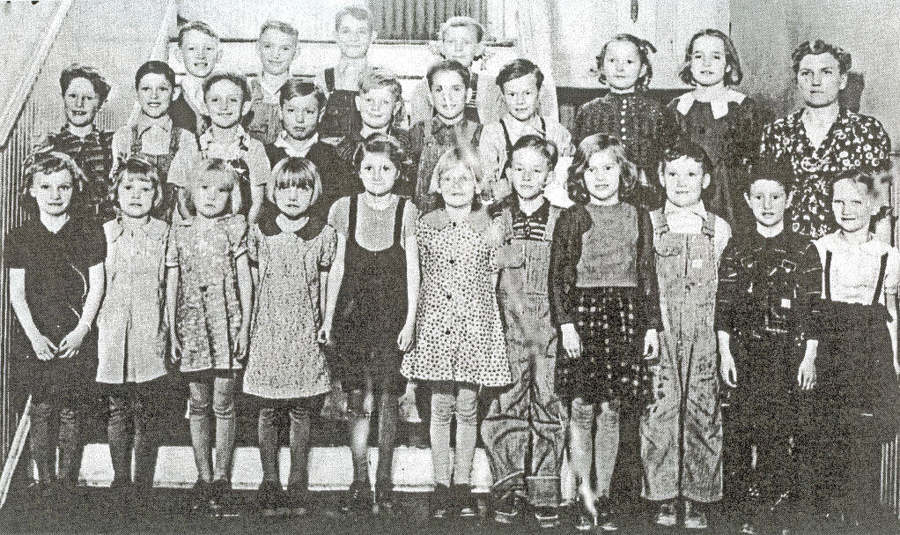 A group of children and a woman, possibly a teacher, stand in rows on a staircase. The children are wearing a variety of clothing styles, including dresses and overalls. The teacher is wearing a patterned dress. All are facing forward, smiling or with neutral expressions. There is no text visible in the image.