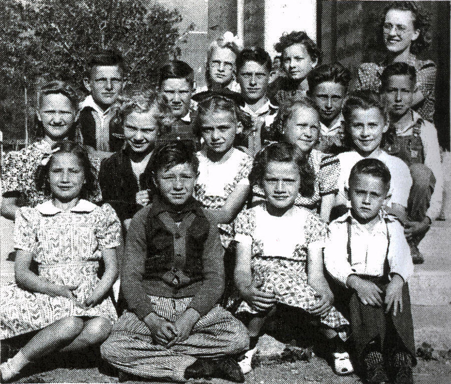 A group of seventeen children, both boys and girls, are seated and standing in rows outside, likely in front of a school building. Most of the children are wearing dresses or shirts with collars, and some have bows in their hair. An adult stands behind the group, smiling. Trees and part of a building are visible in the background.