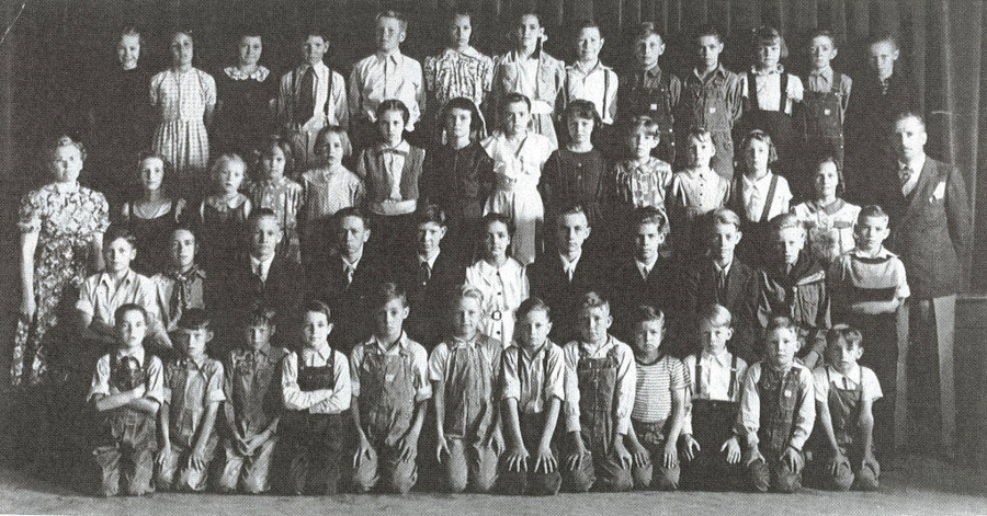 A large group of children, mostly boys, is arranged in multiple rows, with some adults standing on either side. They are wearing a variety of clothing, including overalls and dresses. The setting appears to be indoors with a curtain in the background, suggesting a formal group gathering such as a school class photo.