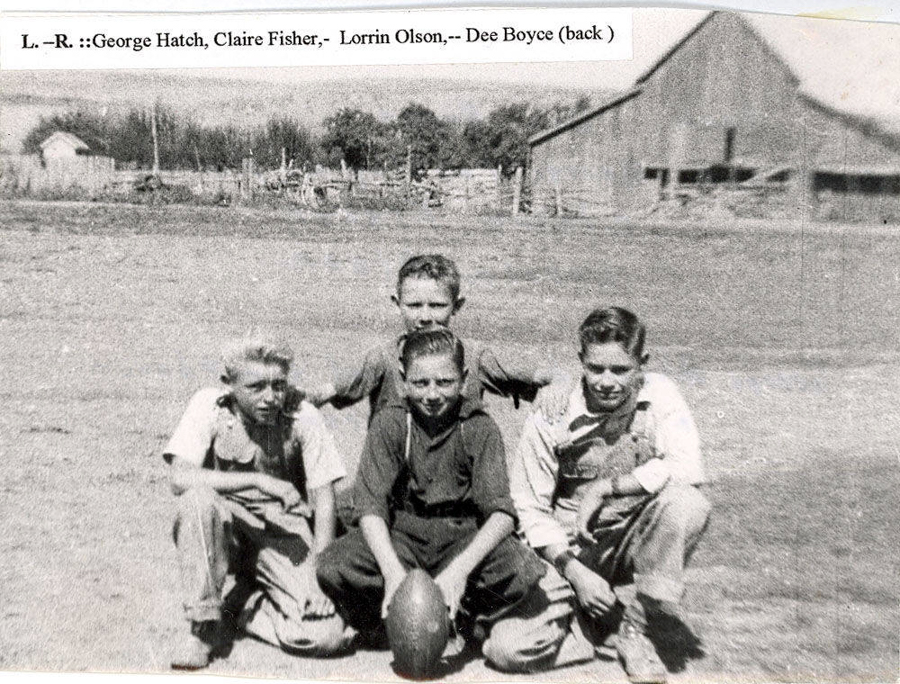 Four boys are sitting and kneeling on a grassy field with a football in front of them. In the background, there is a barn and some trees. There is a label at the top indicating names from left to right: George Hatch, Claire Fisher, Lorrin Olson, and Dee Boyce behind them.