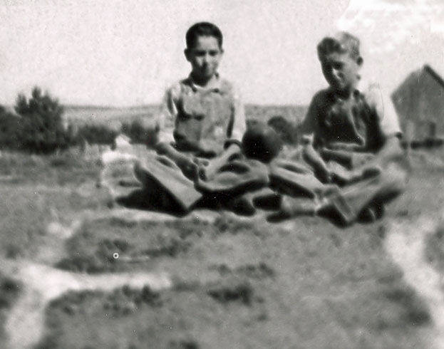 Two boys sitting on the ground in a rural setting. Both are wearing overalls and have their legs crossed. In the background, there is a small building and some vegetation. The scene appears to be outdoors with an open landscape visible.