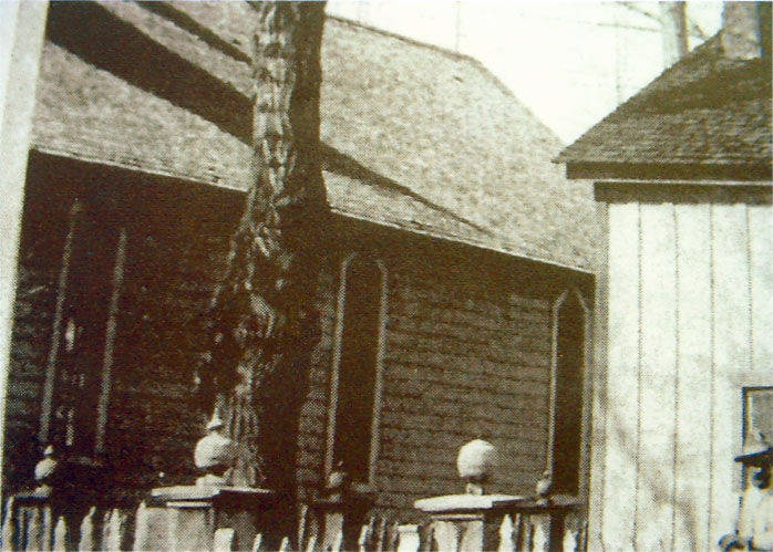 A tall building with arched windows is partially visible, bordered by a wooden fence topped with stone or concrete ornaments. A large tree stands in front of the building, and a smaller structure with a sloped roof is seen on the right side of the image.