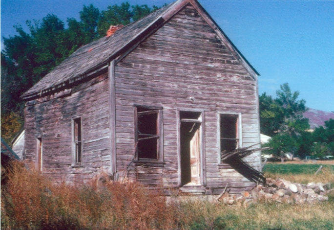 An old, weathered wooden house stands in a grassy area. The structure appears run-down, with a slanted roof and partially broken windows. The front door is slightly ajar, and there is a pile of stones near the foundation. In the background, there are several trees and an open field.