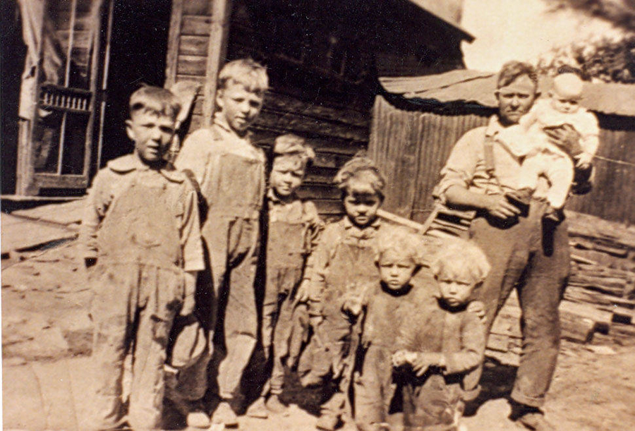 A group of seven children and a man stand outside a rustic wooden building. The children are wearing overalls and appear lined up in front of the man, who holds a baby. The setting looks like a rural area with wooden structures in the background.