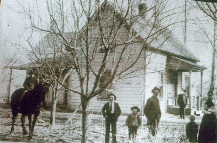 A group of people stand in front of a wooden house with a porch. In the scene, a person is sitting on a horse to the left, and three people, including two adults and one child, are standing in the center. Several other individuals are near the porch of the house. Leafless trees are visible around the area.