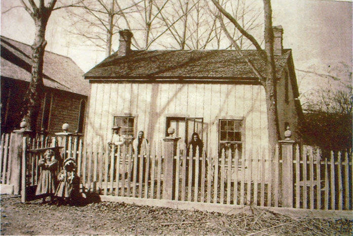 A small wooden house with three windows is surrounded by a picket fence. Six people, including children, stand near or lean on the fence. Two trees flank the house, and the ground is covered with fallen leaves.