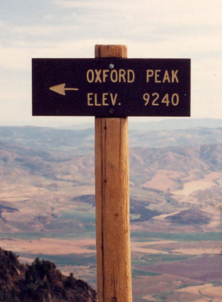 A wooden signpost on a mountain summit with a panoramic view of rolling hills and valleys in the background. The sign reads "Oxford Peak Elev. 9240," with an arrow pointing left.