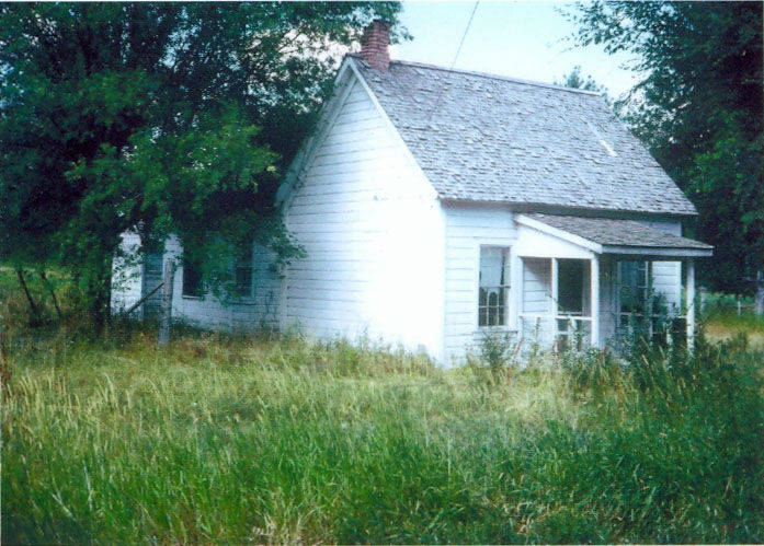 A small, weathered white house with a shingled roof sits in a grassy area surrounded by trees. The house has a chimney and a small porch with two windows. The grass around the house appears overgrown.