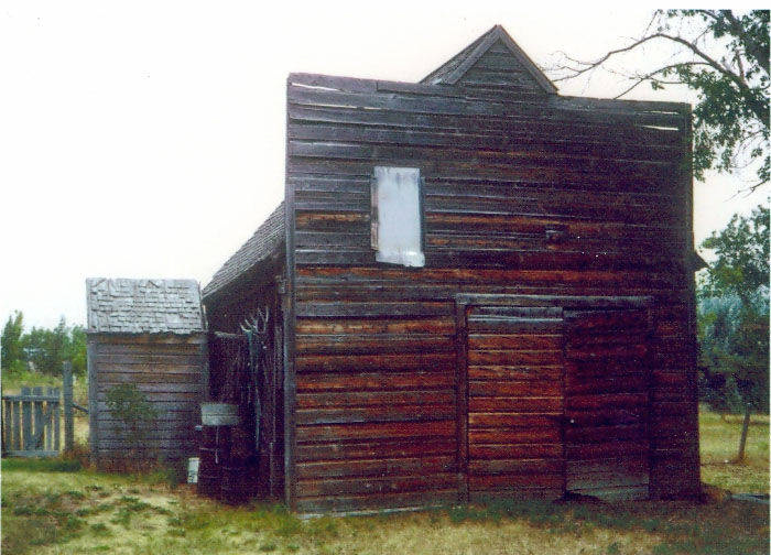 A wooden building with a pitched roof and weathered planks stands on grassy ground. A window is visible on the upper part of the building. To the left, smaller structures with similar construction are attached. Trees and a fence are in the background.