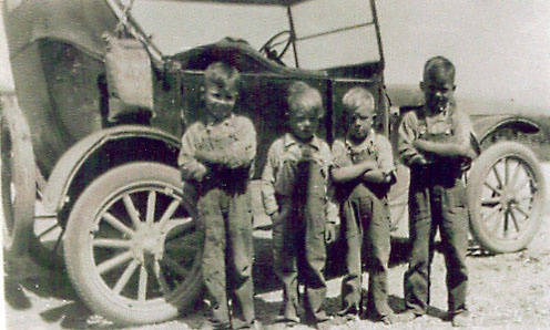 Four children standing in front of an old car. The children are wearing worn clothing and appear to be posing with their arms crossed. The car behind them has large wheels and a folded roof. The scene takes place in an outdoor setting, possibly rural.