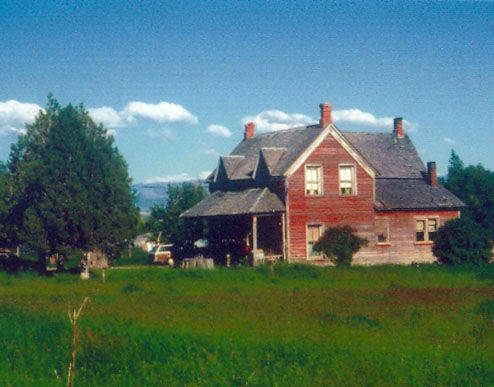 A red wooden house with a steep roof and multiple chimneys, surrounded by trees and situated in a grassy field. There are mountains visible in the distance under a blue sky with clouds.