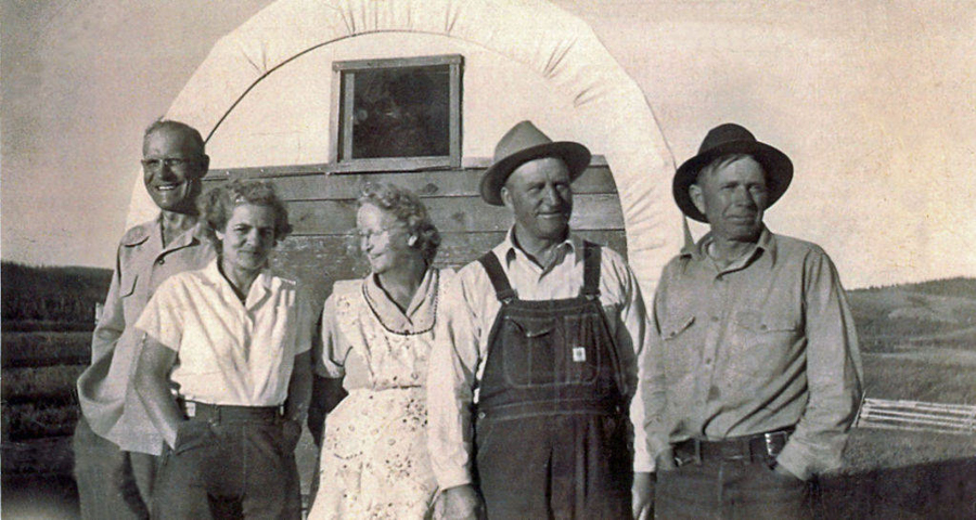 Five people are standing in front of a covered wagon. Three men, two wearing hats, and two women are posed together. They are outdoors, with a wooden fence and open field in the background. The women are wearing dresses, and the men are dressed in shirts and trousers.