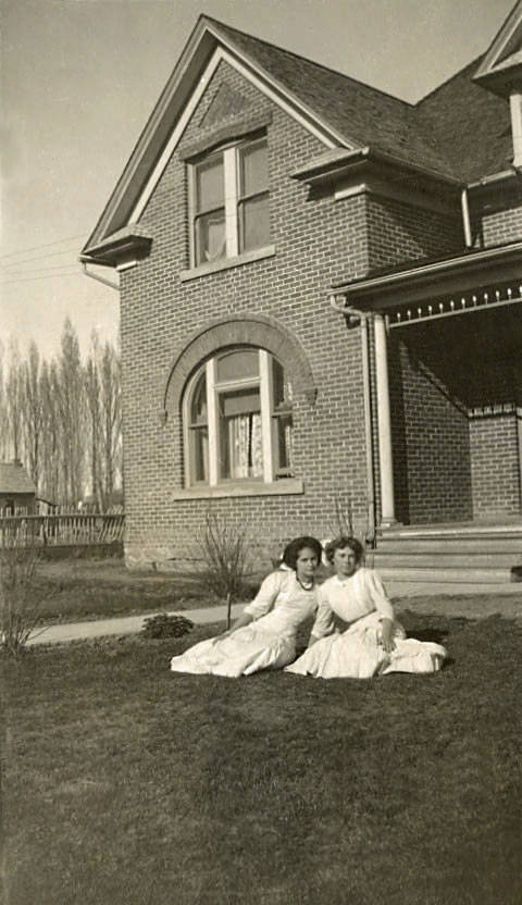 Two women in white dresses are sitting on the grass in front of a large brick house with a triangular roof section, a prominent arched window, and steps leading to a porch. The surrounding yard is landscaped with a few small bushes. In the background, a row of tall trees and a fence are visible.