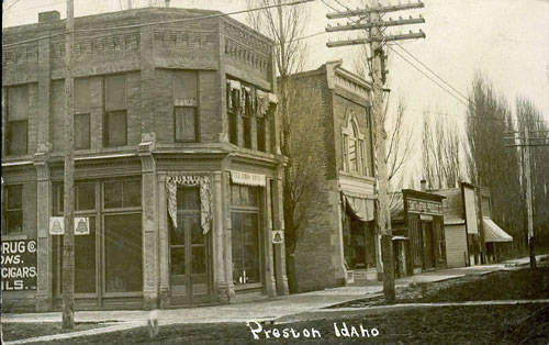 A street scene in a small town with several brick buildings lining the road. The building on the corner has a sign that reads "DRUG", "CIGARS," and "OILS." Utility poles and bare trees are visible along the sidewalk. The text "Preston Idaho" is written at the bottom.