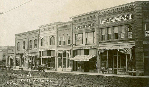 A row of early 20th-century commercial buildings along a street. Businesses include a drugstore, Foss Bros., and John Larson & Sons, with some signs visible above the storefronts. Awning-covered sidewalks and a dirt road run in front. The text "Main St. Preston, Idaho" is written on the street in the foreground.