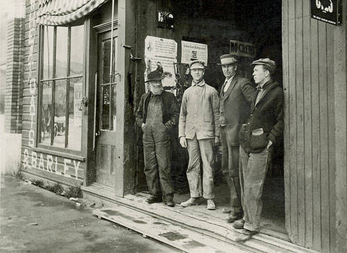 Four men standing in front of the entrance to a building with signage. The first man on the left wears overalls and a cap. The second man is dressed in light-colored work clothes. The third man wears a suit and hat, and the fourth is dressed in a jacket and cap. The words "McGREGOR'S BARLEY" are visible on the window. Posters and signs are displayed on the wall behind them.