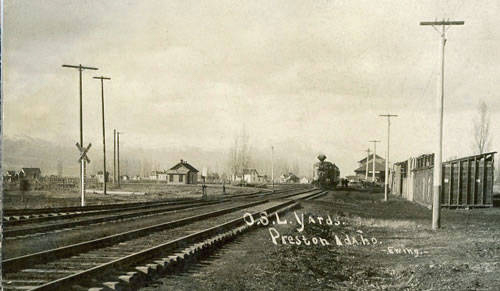 Railroad tracks extend into the distance with a train visible further along the tracks. Several utility poles line the tracks on both sides. A few small buildings and fences are situated near the tracks. In the foreground, text reads "O.S.L. Yards Preston Idaho."