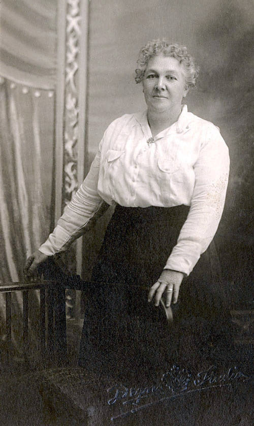 A woman with short, curly hair stands next to a chair. She is wearing a light-colored blouse and a dark skirt. There is ornate drapery in the background. The words "Palace Studio" appear at the bottom.