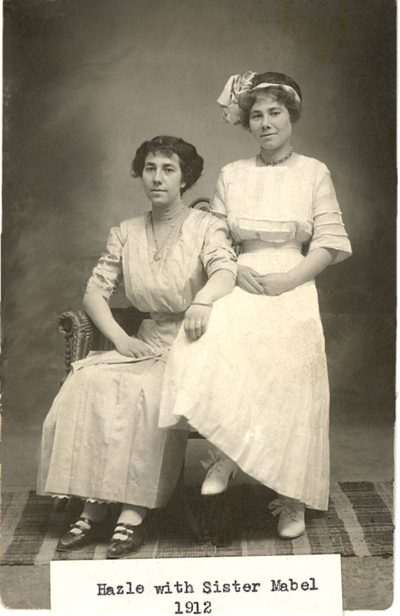 Two women are posed together, with one sitting on a chair and the other sitting on the arm of the chair. Both are wearing long dresses and the woman on the right has a hat with a large bow. A woven mat is on the ground beneath them. There is a label with the text "Hazle with Sister Mabel 1912."