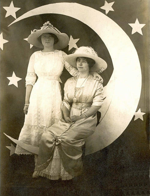 Two women wearing wide-brimmed hats and long dresses are posed on a crescent moon. One woman is sitting while the other stands beside her, resting a hand on her shoulder. The background features several stars.
