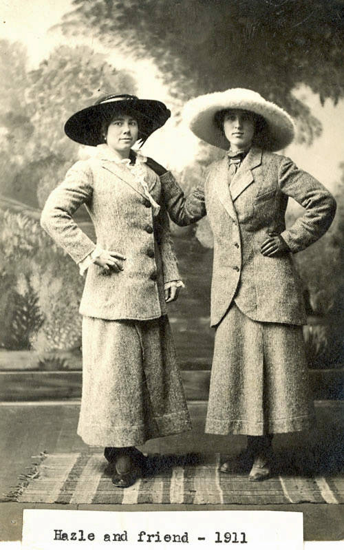 Two women stand side by side on a patterned rug. They are wearing long skirts, tailored jackets, and large hats. One woman has her arm around the other's shoulder. Below them is a label that reads, "Hazle and friend - 1911." Trees are visible in the background.