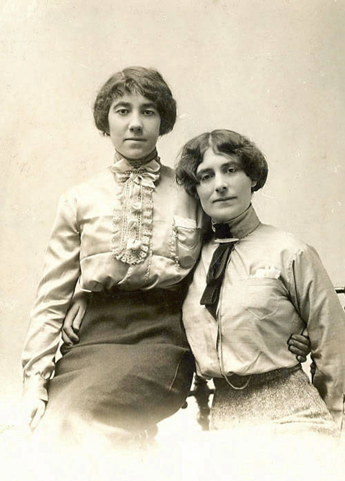 Two women are sitting together. One woman has her arm around the other. They are both wearing long-sleeved blouses and skirts. The woman on the left is wearing a blouse with a ruffled front, while the woman on the right has a bow tie on her blouse.