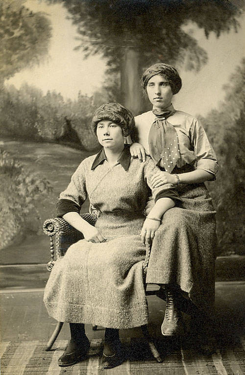 Two women are posed in front of a painted backdrop depicting a natural scene. One woman is seated in a chair, wearing a dress with dark trim on the cuffs, and looking slightly to the side. The other woman stands beside her, resting a hand on the seated woman's shoulder, wearing a blouse with a polka-dot necktie and a skirt. Both women wear head coverings and boots.