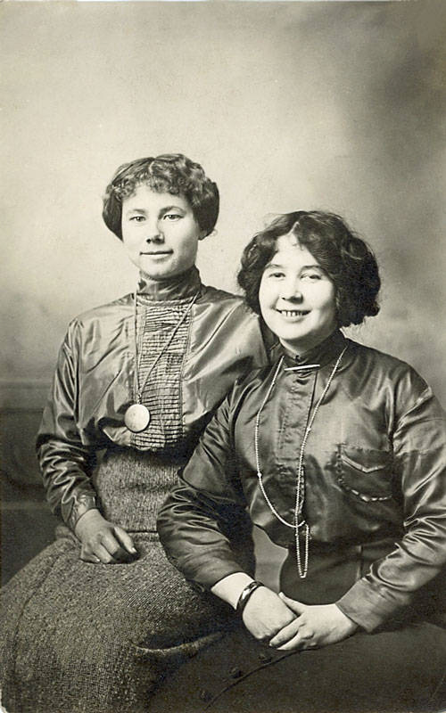 Two women are seated side by side. The woman on the left has short, wavy hair and wears a high-necked blouse with a long necklace and pendant. The woman on the right has curly hair and smiles, wearing a blouse with a chain necklace and a wrist bracelet. Their attire appears formal.