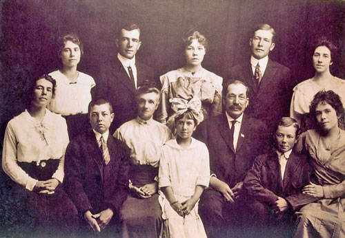 A group of thirteen people is posed for a portrait. Four men and three women stand in the back row. In the front row, two women, two young boys, one young girl, and one man are seated. The young girl in the center front row is wearing a large decorative hat. The man seated next to the girl is wearing a suit and tie, with a pin on his lapel. The people are arranged closely together, with neutral expressions.