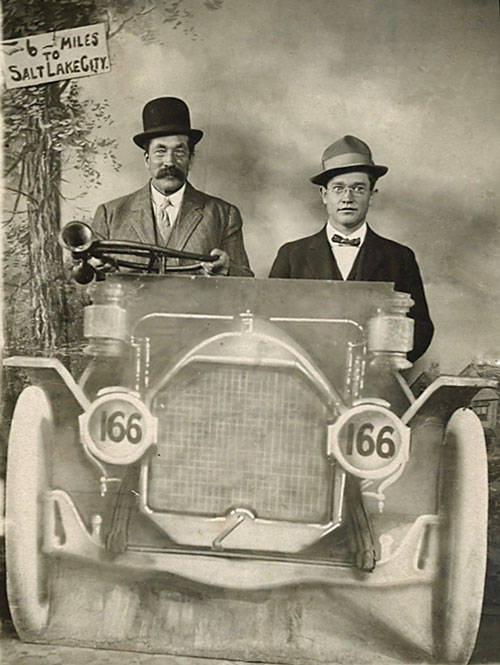 Two men are seated in an early automobile, with the number 166 visible on the front. The man on the left holds a horn. Both men are wearing suits and hats. In the background, there is a painted backdrop with a sign that reads "6 to 6 Miles to Salt Lake City."