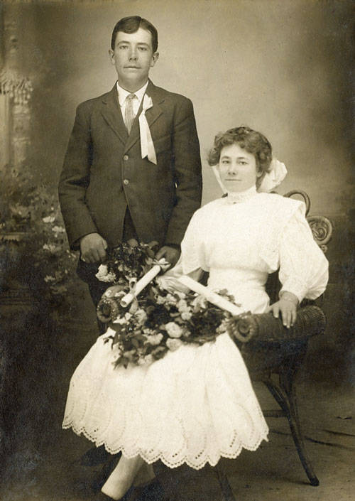 A man and a woman pose for a portrait. The man stands beside the woman, wearing a dark suit with a light tie and a ribbon on his chest. The woman sits in an ornate chair, dressed in a long white dress, holding a large bouquet of flowers. The setting has a formal, vintage studio backdrop with floral decorations.