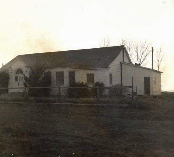 A single-story building with a pitched roof, featuring a large arched window and several rectangular windows. It is surrounded by a simple wooden fence and has a few shrubs and bare trees nearby. The structure resembles a house or small community building.