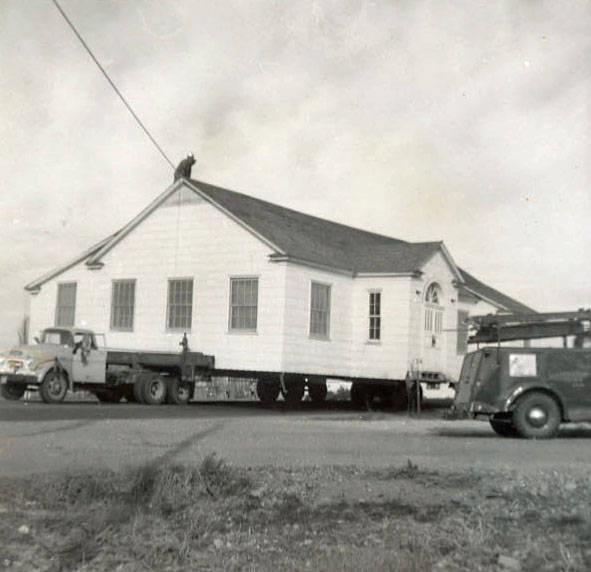 A one-story building is being transported on the back of a large flatbed truck, with a person visible on the roof. Another vehicle is parked nearby, featuring text on its side, but the text is not clear. The scene takes place at an intersection with some visible plants in the foreground.