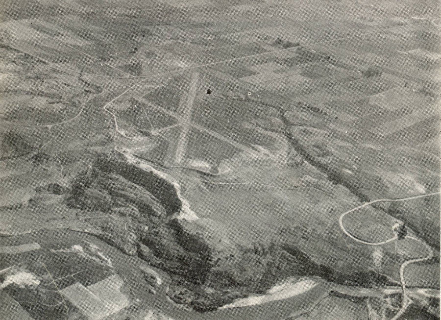 Aerial view of a landscape featuring a runway, several winding roads, and a river. The surrounding area includes a patchwork of fields and open land. The scene is expansive, with no text visible.