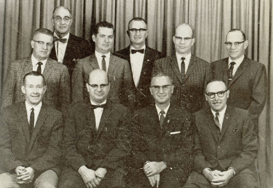 A group of ten men in suits pose together in front of a curtain. Some are wearing glasses and most have ties. The men are arranged in three rows, with four standing in the back, three in the middle, and three seated in the front.