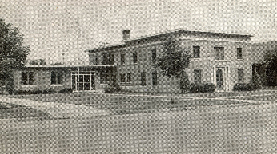 A two-story brick building with a rectangular structure, surrounded by neatly trimmed shrubs and small trees. The entrance features a decorative arch above the doorway. The building is set back from the street with a lawn and sidewalks leading up to it. There are power lines visible in the background.