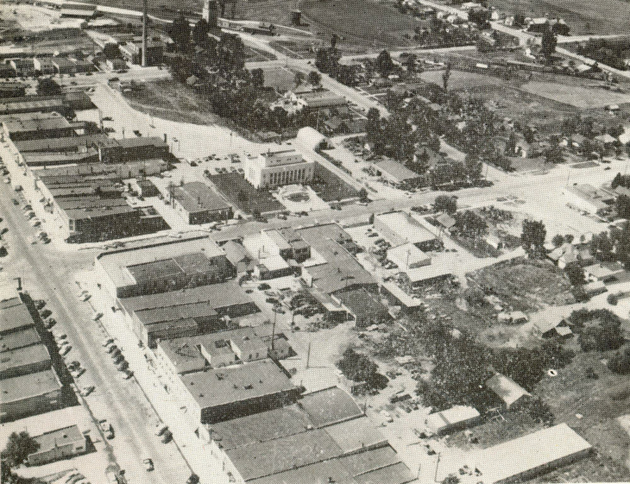 An aerial view of a small town with a grid layout of streets and buildings. There are several one-story structures, likely shops or houses, and a few parked cars along the streets. Trees and small patches of greenery are scattered between the buildings. There is a larger, more prominent building near the center, possibly a public or municipal structure. In the background, expansive open land is visible.