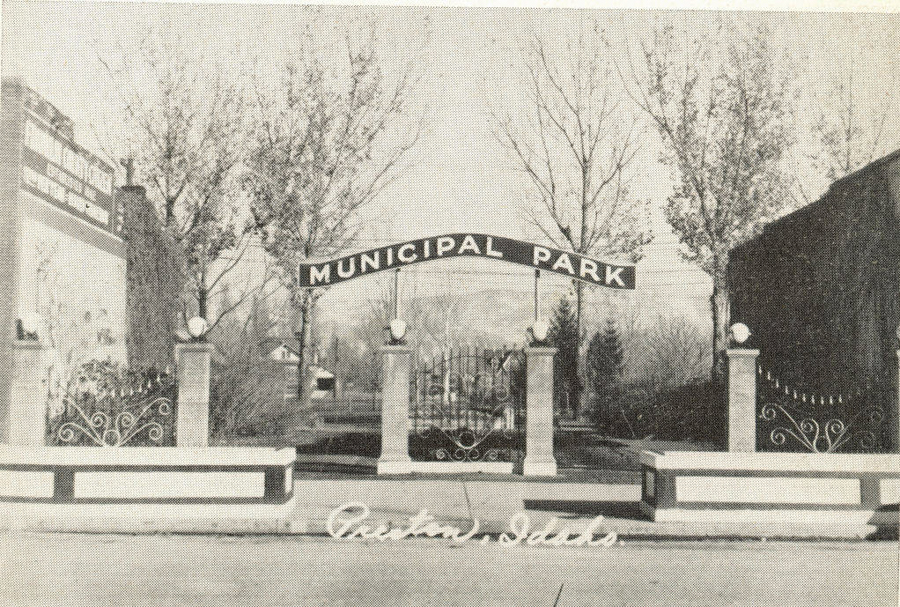 A decorative iron gate with stone pillars stands in front of a park. A sign above the entrance reads "Municipal Park." There are trees in the background. The word "Cedar City" appears at the bottom.