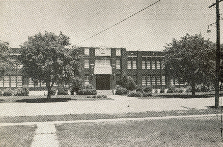 A large brick building with two rows of windows and a central entrance. Trees and shrubs are in front of the building. A sidewalk leads to the entrance, and there is a telephone pole and power line in the foreground.