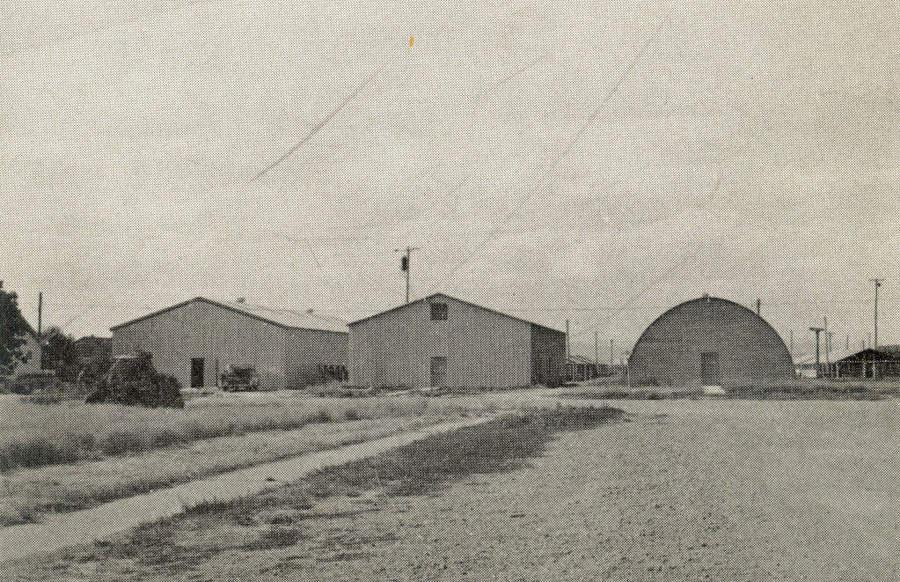 A landscape with three large buildings. The first two are rectangular with pitched roofs, and the third has a rounded, Quonset hut shape. The buildings are surrounded by open, grassy fields with a dirt path in the foreground. Power poles and a few scattered objects are visible around the buildings.