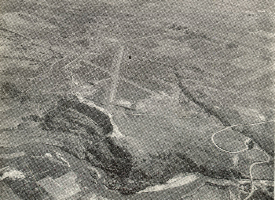 Aerial view of a landscape showing a large expanse of farmland divided into rectangular plots. A river winds through the lower part of the scene. There are visible roads and a runway crossing the fields. Sparse vegetation and gentle hills are scattered throughout the area.