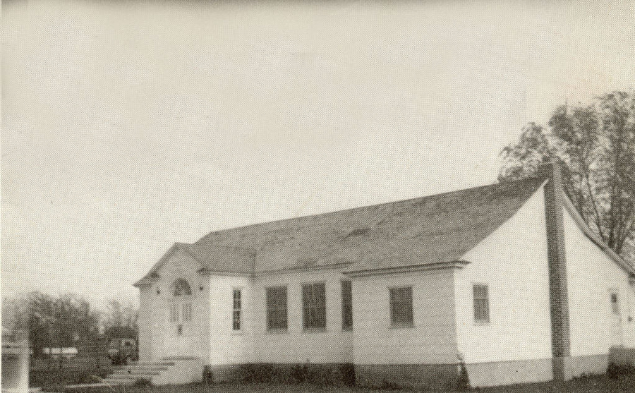 A small, single-story building with a sloped roof and arched windows. It has a chimney on one side. There are trees and a vehicle in the background, and a staircase leads to the entrance on the left side.