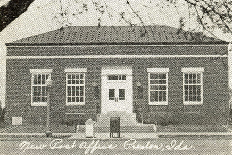 A brick building with a flat roof and a symmetrical facade featuring a central entrance flanked by large windows. The words "UNITED STATES POST OFFICE" are written above the entrance. A lamppost and a mailbox are situated in front of the building. The text "New Post Office Preston, Ida." is written across the bottom. Tree branches are visible in the foreground.