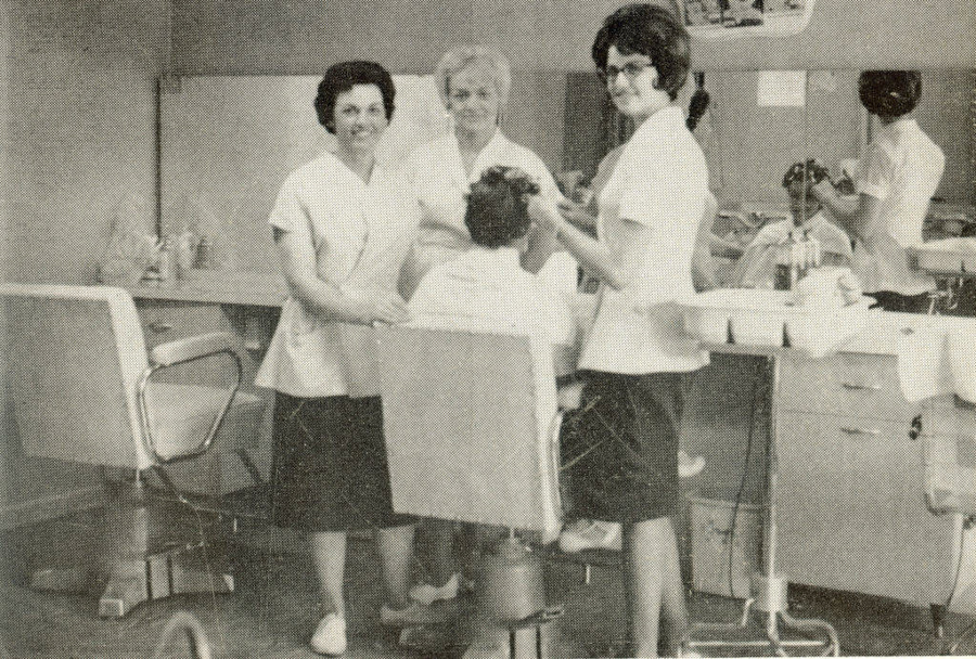 Three women in uniform are standing around a seated person in a beauty salon. One is styling the seated person's hair, while the others are observing. The salon has styling chairs, mirrors, and various hair products and tools on the counter. Another stylist is visible in the background working on a client.