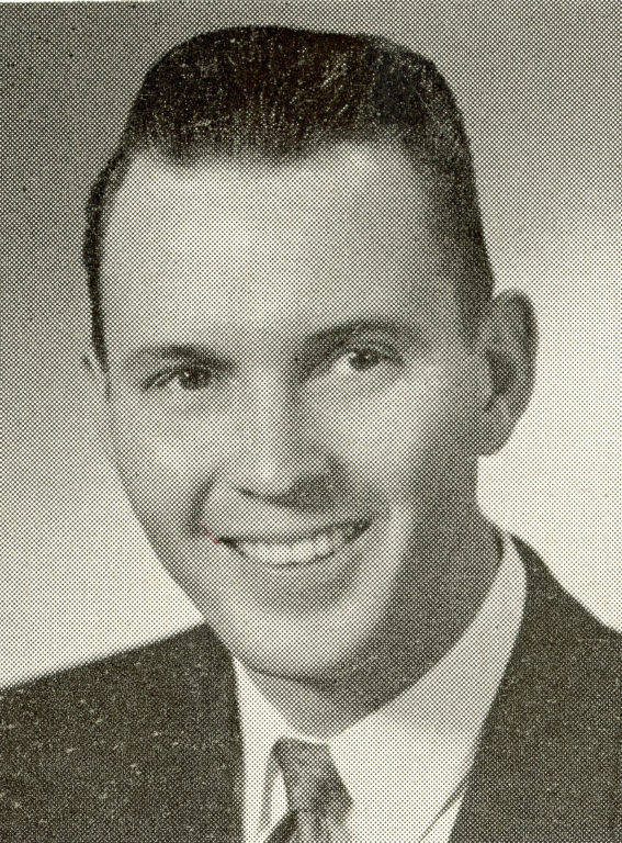 A man is smiling and wearing a suit with a tie. He has short hair that is styled neatly. The background is plain. There is no text in the image.