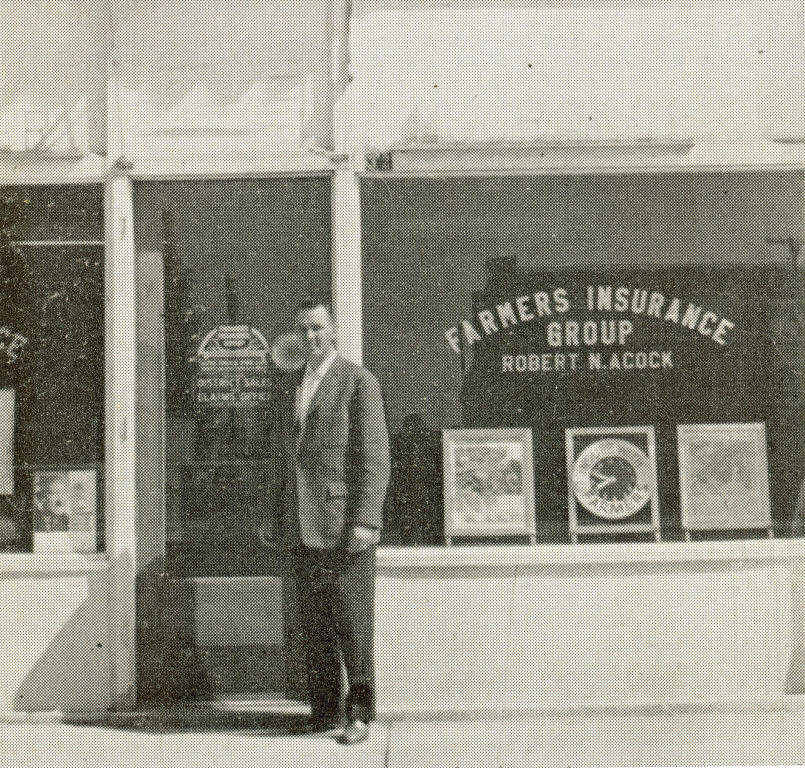 A man in a suit stands in front of a storefront. The window displays the text "Farmers Insurance Group Robert W. Acock." Signs and posters are visible inside the window.