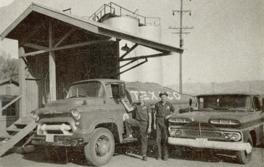 Two men in uniforms stand in front of a vintage truck and a pickup. They are at a structure with a slanted roof and stairs. Behind them, a large tank with the word "TEXACO" is visible, alongside a telephone pole.